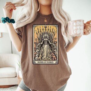 May include: Brown t-shirt featuring a tarot card-style graphic. The graphic depicts a woman on a throne surrounded by poodles, with the text "MOTHER OF POODLES" at the bottom. The design is in shades of yellow, beige, and brown.