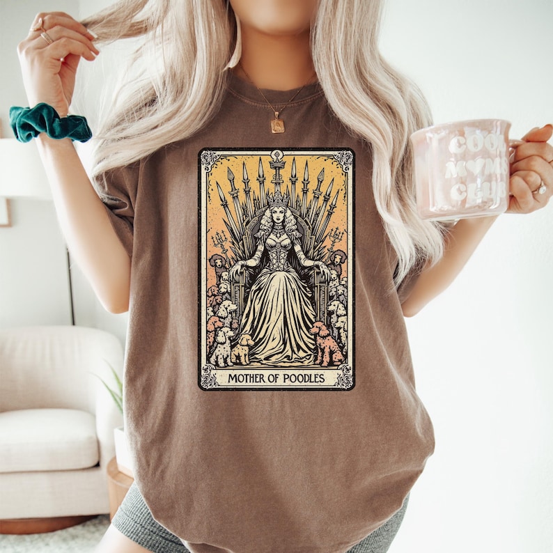 May include: Brown t-shirt featuring a tarot card-style graphic. The graphic depicts a woman on a throne surrounded by poodles, with the text "MOTHER OF POODLES" at the bottom. The design is in shades of yellow, beige, and brown.