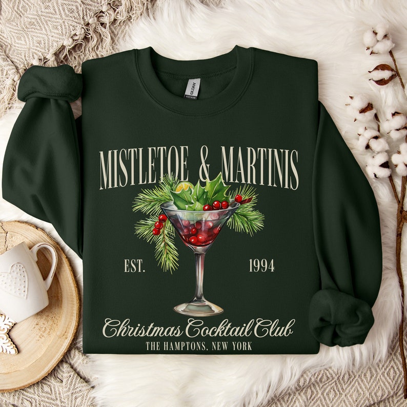 May include: A dark green long-sleeve sweatshirt featuring a graphic of a martini glass filled with red berries and pine needles. The text reads MISTLETOE & MARTINIS, EST. 1994, Christmas Cocktail Club, and THE HAMPTONS, NEW YORK in a cream-colored font.