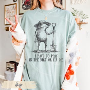 May include: A light blue t-shirt features a whimsical illustration of a frog wearing a hat and sunglasses, holding a shovel. The text "I HAVE TO PLAY IN THE DIRT OR I'LL DIE" is printed below the frog. The shirt is a relaxed fit.