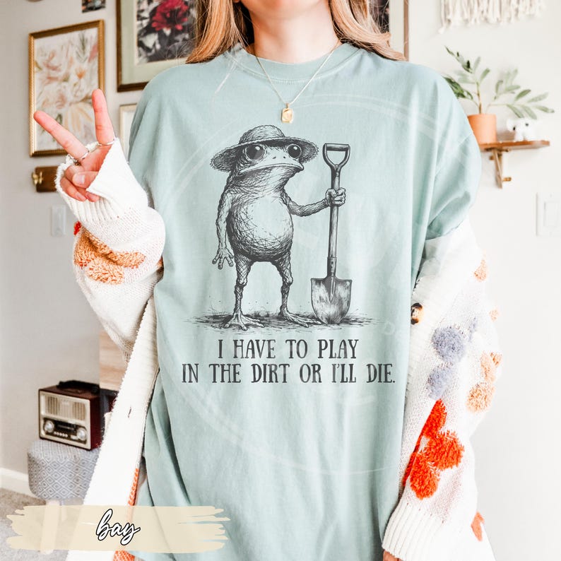 May include: A light blue t-shirt features a whimsical illustration of a frog wearing a hat and sunglasses, holding a shovel. The text "I HAVE TO PLAY IN THE DIRT OR I'LL DIE" is printed below the frog. The shirt is a relaxed fit.