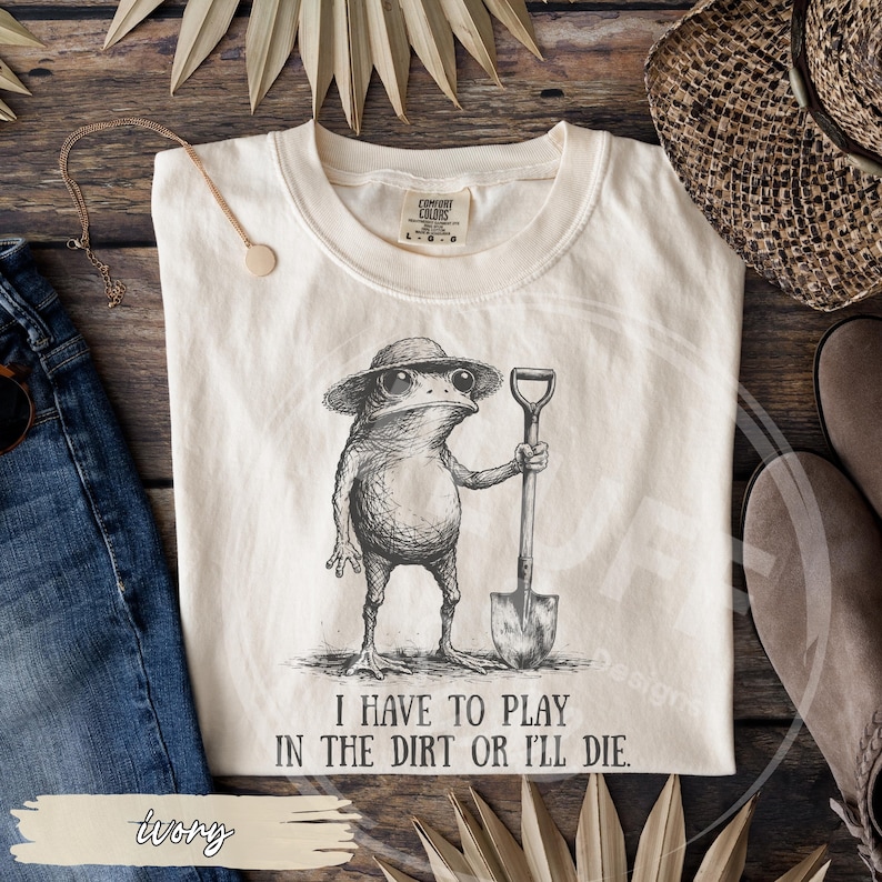 May include: Ivory-colored t-shirt featuring a detailed illustration of a frog wearing a hat and holding a shovel. The text "I HAVE TO PLAY IN THE DIRT OR I'LL DIE" is printed below the frog. The shirt is a Comfort Colors brand.