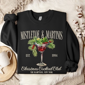 May include: A black crewneck sweatshirt featuring a graphic of a martini glass filled with red liquid, garnished with green pine needles, holly leaves, and red berries. The text reads MISTLETOE & MARTINIS, EST. 1994, Christmas Cocktail Club, THE HAMPTONS, NEW YORK.