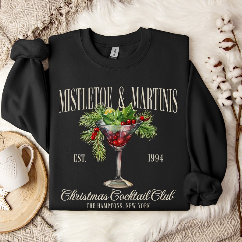 May include: A black crewneck sweatshirt featuring a graphic of a martini glass filled with red liquid, garnished with green pine needles, holly leaves, and red berries. The text reads MISTLETOE & MARTINIS, EST. 1994, Christmas Cocktail Club, THE HAMPTONS, NEW YORK.