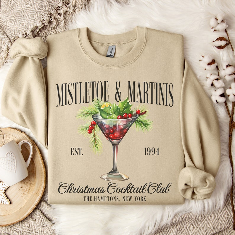 May include: A beige crewneck sweatshirt featuring a graphic of a martini glass filled with red berries and green holly leaves. The text reads MISTLETOE & MARTINIS, EST. 1994, Christmas Cocktail Club, and THE HAMPTONS, NEW YORK in black lettering.