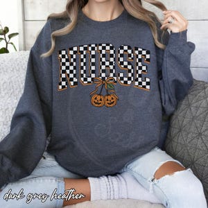 Puede incluir: Una sudadera gris jaspeado oscuro con la palabra NURSE en un patr&oacute;n de cuadros blanco y negro. Debajo del texto, dos calabazas naranjas con caras talladas est&aacute;n atadas con un lazo, parecidas a cerezas, con una peque&ntilde;a hoja verde.
