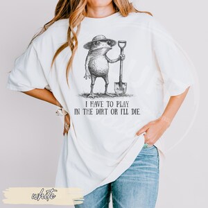 May include: White t-shirt featuring a whimsical illustration of a frog wearing a hat and holding a shovel. The text "I HAVE TO PLAY IN THE DIRT OR I'LL DIE" is printed below the frog. The shirt is a relaxed fit.