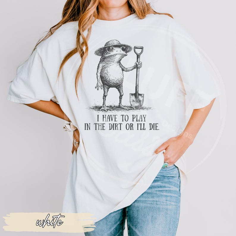 May include: White t-shirt featuring a whimsical illustration of a frog wearing a hat and holding a shovel. The text "I HAVE TO PLAY IN THE DIRT OR I'LL DIE" is printed below the frog. The shirt is a relaxed fit.