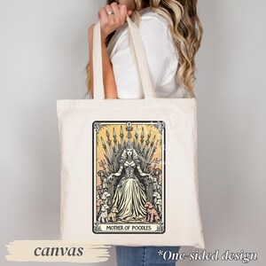 May include: Cream-colored canvas tote bag featuring a tarot card-style illustration. The design depicts a woman on a throne surrounded by poodles, with the text "MOTHER OF POODLES." The bag has long handles and the word "canvas" is printed on the bottom.