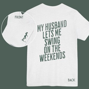 Może przedstawiać: Biała koszulka z nadrukiem "My husband lets me swing on the weekends" na plecach w ciemnozielonym kolorze. Prz&oacute;d koszulki ma mały grafik przedstawiający kobietę na nartach wodnych.