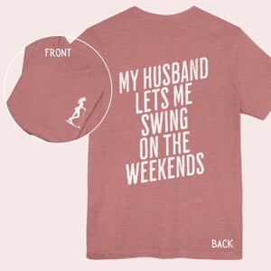 Może przedstawiać: R&oacute;żowy t-shirt z nadrukiem "My husband lets me swing on the weekends" na plecach w kolorze białym.