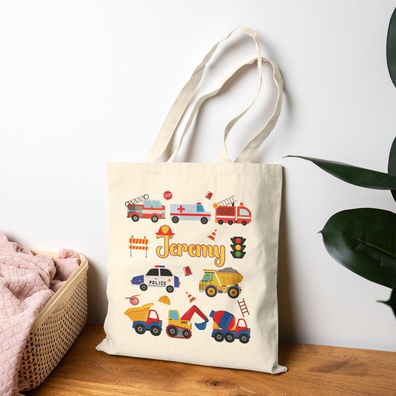 Puede incluir: Bolso tote de color crema con ilustraciones coloridas de veh&iacute;culos de emergencia y equipos de construcci&oacute;n. El nombre "Jeremy" est&aacute; impreso en una fuente divertida. Incluye camiones de bomberos, una ambulancia, un coche de polic&iacute;a y un cami&oacute;n volquete.