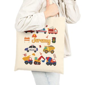 Puede incluir: Bolso tote color crema con ilustraciones coloridas de veh&iacute;culos de emergencia y equipos de construcci&oacute;n. El nombre "Jeremy" est&aacute; impreso en amarillo. Los dise&ntilde;os incluyen un cami&oacute;n de bomberos, un coche de polic&iacute;a, un cami&oacute;n volquete y sem&aacute;foros. El bolso lo sostiene una persona.