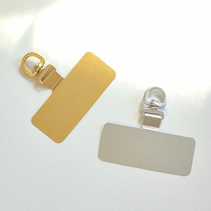 May include: Two metal phone charms. One is gold-colored, the other is silver-colored. Each charm has a rectangular plate with a swivel ring and a clasp for attaching to a phone case or other item. The charms are shown on a white background.