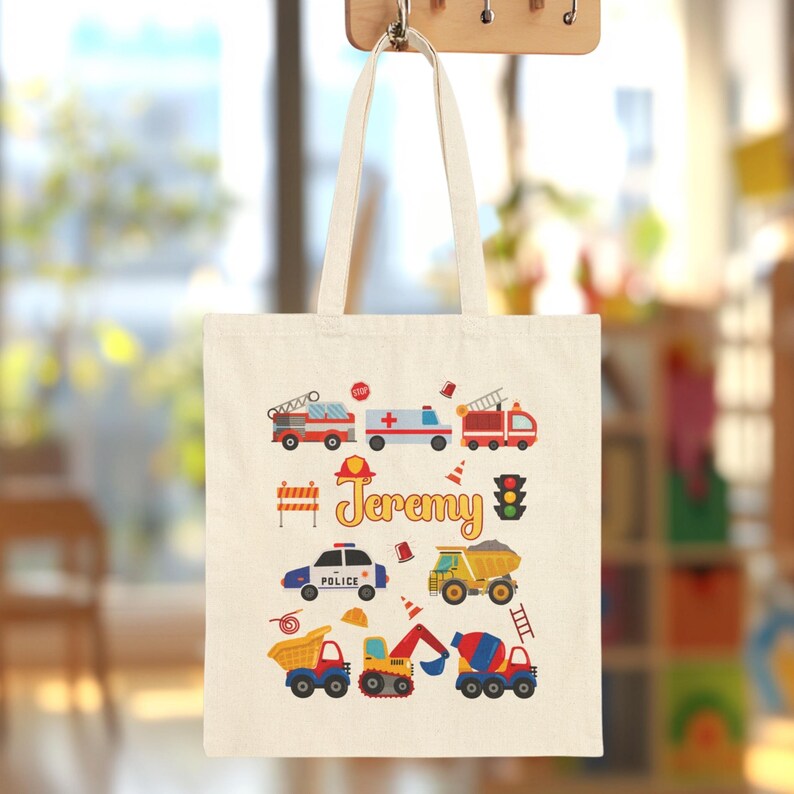 Puede incluir: Bolso tote de color crema con ilustraciones coloridas de veh&iacute;culos de emergencia y equipos de construcci&oacute;n. El nombre "Jeremy" est&aacute; impreso en escritura amarilla. Incluye camiones de bomberos, una ambulancia, un coche de polic&iacute;a, volquetes y una hormigonera.