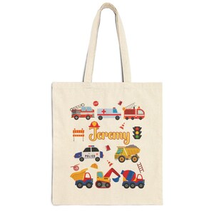 Puede incluir: Una bolsa de mano de lona beige con ilustraciones coloridas de veh&iacute;culos de emergencia y equipos de construcci&oacute;n. El nombre "Jeremy" est&aacute; escrito en amarillo. Incluye un cami&oacute;n de bomberos, una ambulancia, un coche de polic&iacute;a y volquetes.