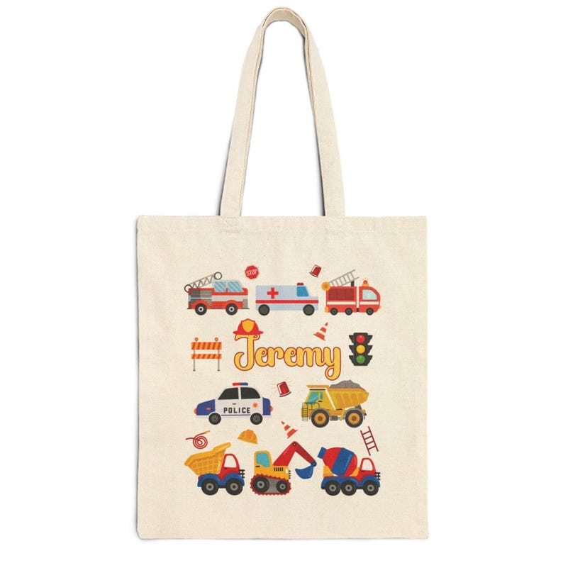 Puede incluir: Una bolsa de mano de lona beige con ilustraciones coloridas de veh&iacute;culos de emergencia y equipos de construcci&oacute;n. El nombre "Jeremy" est&aacute; escrito en amarillo. Incluye un cami&oacute;n de bomberos, una ambulancia, un coche de polic&iacute;a y volquetes.