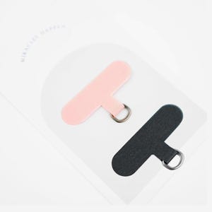 May include: Two phone charms, one pink and one black, each with a silver-colored ring. The charms are on a white background with the words "MIRACLES HAPPEN" in light gray.