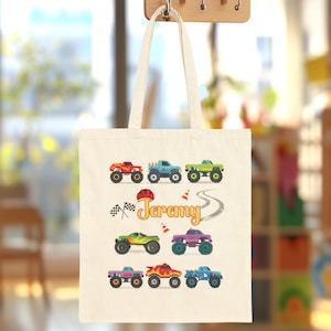 May include: A cream-colored tote bag featuring colorful monster truck illustrations. The bag has the name "Jeremy" in a playful font, along with racing flags and a tire track design. The bag is hanging on a wooden hook.