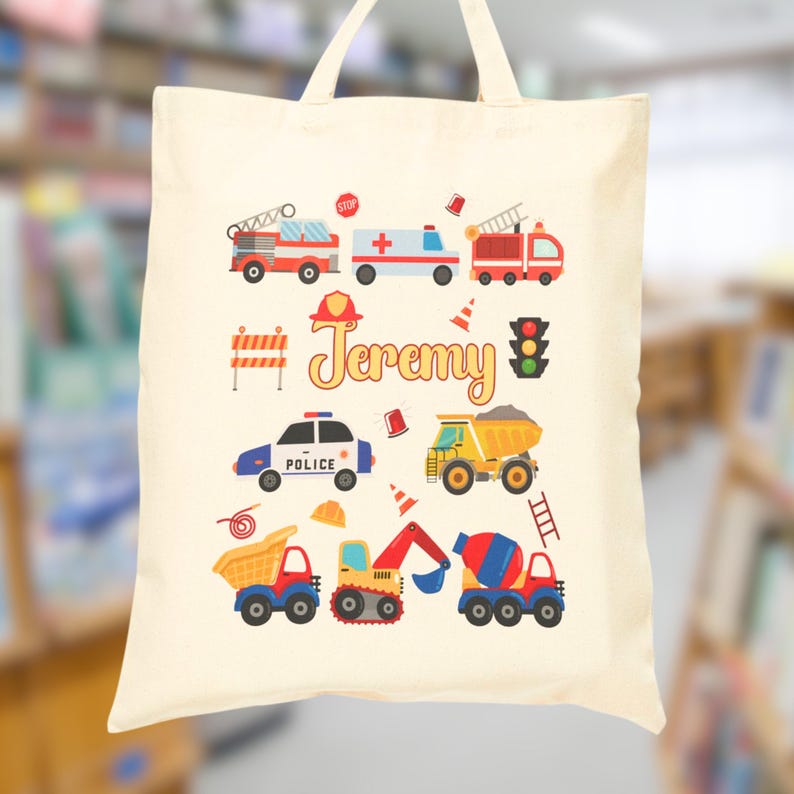 Puede incluir: Un bolso tote de color crema con ilustraciones coloridas de veh&iacute;culos de emergencia y equipos de construcci&oacute;n. El nombre "Jeremy" est&aacute; impreso en una fuente l&uacute;dica. Incluye un cami&oacute;n de bomberos, una ambulancia, un coche de polic&iacute;a y volquetes.