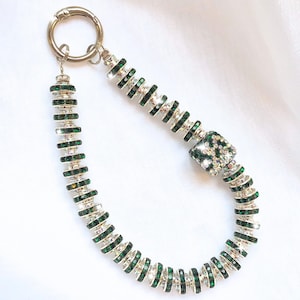 May include: A silver-toned keychain with a series of green and clear crystal accents. The keychain features a circular clasp and a decorative chain with a square crystal centerpiece. The crystals are arranged in a circular pattern.