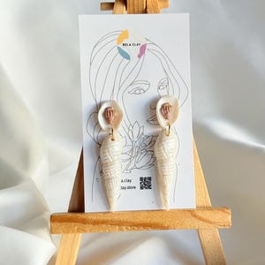 Peut inclure: Boucles d'oreilles en forme de coquillage, présentées sur un chevalet en bois. Les boucles d'oreilles sont blanches, avec une surface texturée et un détail rose doré. Le fond présente une carte blanche avec un dessin au trait et le texte "Bela Clay".