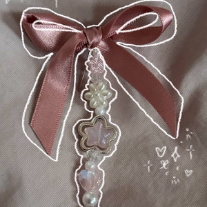 May include: A pink satin bow with a Hello Kitty charm hanging from it. The charm is white with pink accents and a yellow face. The bow is decorated with white and pink beads and a star-shaped charm.