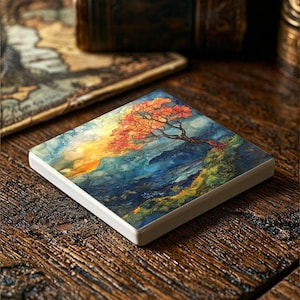 May include: A square ceramic coaster with a watercolor painting of a tree with orange leaves against a blue and green mountain landscape.