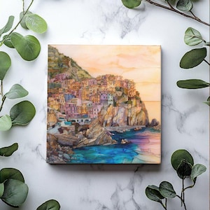 May include: A watercolor painting of a colorful Italian village built on a cliffside overlooking a turquoise blue ocean. The sky is a soft orange and pink.