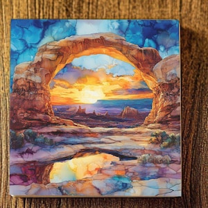 May include: A watercolor painting of a natural sandstone arch in a desert landscape. The arch is illuminated by a bright orange sunset, casting long shadows across the scene. The sky is a vibrant blue with wispy clouds. A small pool of water reflects the sunset colors.