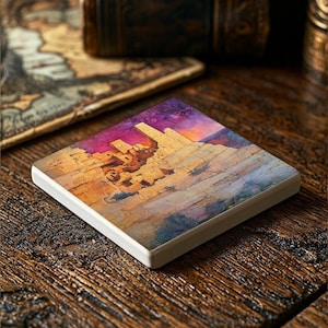 May include: A square ceramic coaster with a watercolor painting of a cliffside village with a purple and pink sky. The village is made of stone and has a few buildings.