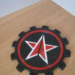 May include: A black and red 3D printed star with a white outline, set against a black background. The star is surrounded by a black gear-like ring with 12 points.