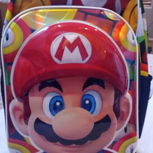May include: A red and blue backpack featuring a large image of Mario from the Super Mario Bros. video game series. The backpack has a white zip and a white trim around the edges.