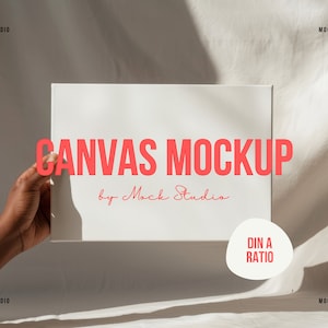 Puede incluir: Una maqueta de lienzo blanco sostenida por una mano sobre un fondo de tela blanca. El lienzo tiene el texto "CANVAS MOCKUP by Mock Studio" impreso en rojo. Un círculo blanco con el texto "DIN A RATIO" impreso en rojo se encuentra en la esquina inferior derecha de la imagen.