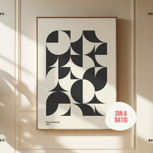 K&ouml;nnte beinhalten: Ein gerahmter Poster-Mockup mit einem Schwarz-Wei&szlig;-Druck eines geometrischen abstrakten Kunstwerks mit dem Text "BAUHAUS 2023". Das Poster h&auml;ngt an einer wei&szlig;en Wand mit einer hellbraunen Holzleiste. Der Text "DIN A RATIO" ist in Rot auf einem wei&szlig;en Kreis geschrieben.