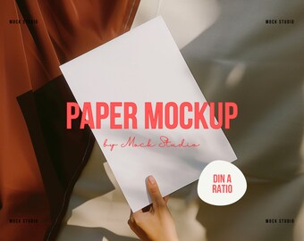 Puede incluir: Una hoja de papel blanca sostenida por una mano sobre un fondo de tela marrón y beige. El papel está en blanco y tiene el texto "PAPER MOCKUP by Mock Studio" y "DIN A RATIO" escrito en él.