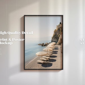 May include: A framed print mockup of a beach scene with white umbrellas and lounge chairs on a sandy beach. The beach is next to a rocky cliff with a small town on top. The print is in a black frame and is hanging on a white wall. The text "A1 A3" is visible in the upper right corner of the image.