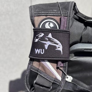 May include: Black scuba diving backpack with a white shark logo and the letters "WU" on a black strap.