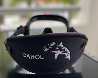 May include: A black neoprene diving mask strap cover featuring an embroidered white hammerhead shark graphic and the name CAROL stitched in white thread. The cover has a curved shape and includes black adjustable side straps for secure attachment.