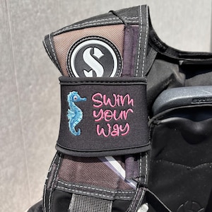 Op de afbeelding: Zwarte duikuitrusting met een zeepaardje en de tekst "Swim your Way" geborduurd in roze. De uitrusting heeft ook een wit en zwart logo.