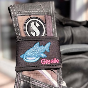 Puede incluir: Una bolsa negra de equipo de buceo con un parche de tiburón ballena azul y el nombre "Giselle" bordado en rosa.