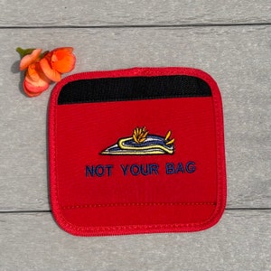 Puede incluir: Posavasos de tela roja con ribete negro y una babosa marina bordada en azul y amarillo con el texto "NOT YOUR BAG" en azul.