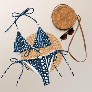 May include: A two-piece bikini set with a navy blue and white pattern. The triangle top and tie-side bottoms are displayed on a woven mat, alongside a round straw bag and sunglasses. The bikini features a repeating dot and stripe design.