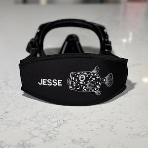 Puede incluir: Máscara de buceo negra con lentes transparentes. La máscara presenta una correa negra con el nombre "JESSE" bordado en blanco, junto con un diseño de pez bordado en blanco. Diseñada para actividades subacuáticas.