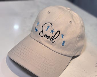 May include: A beige baseball cap with the word "Soeil" embroidered in black script, accented with small blue sea-themed designs. The cap has a curved brim and a classic six-panel design.
