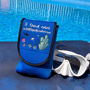 May include: A blue scuba mask case with the text "If found email info@liquidscuba.com" and marine life illustrations. A white scuba mask rests beside the case against a blue surface, with a swimming pool in the background.