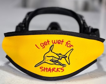 May include: A black scuba diving mask featuring a bright yellow neoprene strap cover. The cover is embroidered with a black shark illustration and the red text "I get wet for SHARKS" centered on the front surface of the yellow fabric.