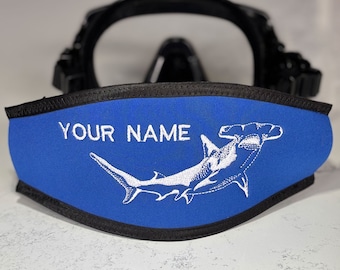 May include: A blue neoprene scuba mask strap cover with black trim, featuring an embroidered white hammerhead shark and the text "YOUR NAME" in white capital letters, positioned in front of a black scuba diving mask frame.