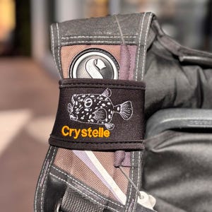 Puede incluir: Una mochila negra y gris con un pez globo bordado en blanco y negro y el nombre "Crystalle" en hilo naranja.