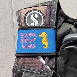 Könnte beinhalten: Ein Tauch-Jacket (BCD) mit einem blauen Band mit dem gestickten Schriftzug "Swim your Way" und einem gelben Seepferdchen. Das BCD hat ein schwarz-braunes Farbschema mit einem schwarz-weißen Logo.